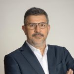 A person wearing a dark blazer over a white dress shirt stands with arms crossed against a plain light-colored background. The individual has short dark hair and is wearing a watch on their left wrist.