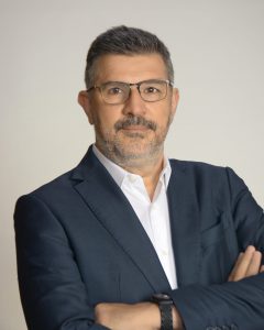 A person wearing a dark blazer over a white dress shirt stands with arms crossed against a plain light-colored background. The individual has short dark hair and is wearing a watch on their left wrist.