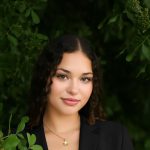 A person wearing a black blazer and a light-colored top stands outdoors surrounded by lush green leaves and branches. They are holding a small branch in one hand and wearing two thin necklaces with small gold pendants.