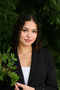 A person wearing a black blazer and a light-colored top stands outdoors surrounded by lush green leaves and branches. They are holding a small branch in one hand and wearing two thin necklaces with small gold pendants.